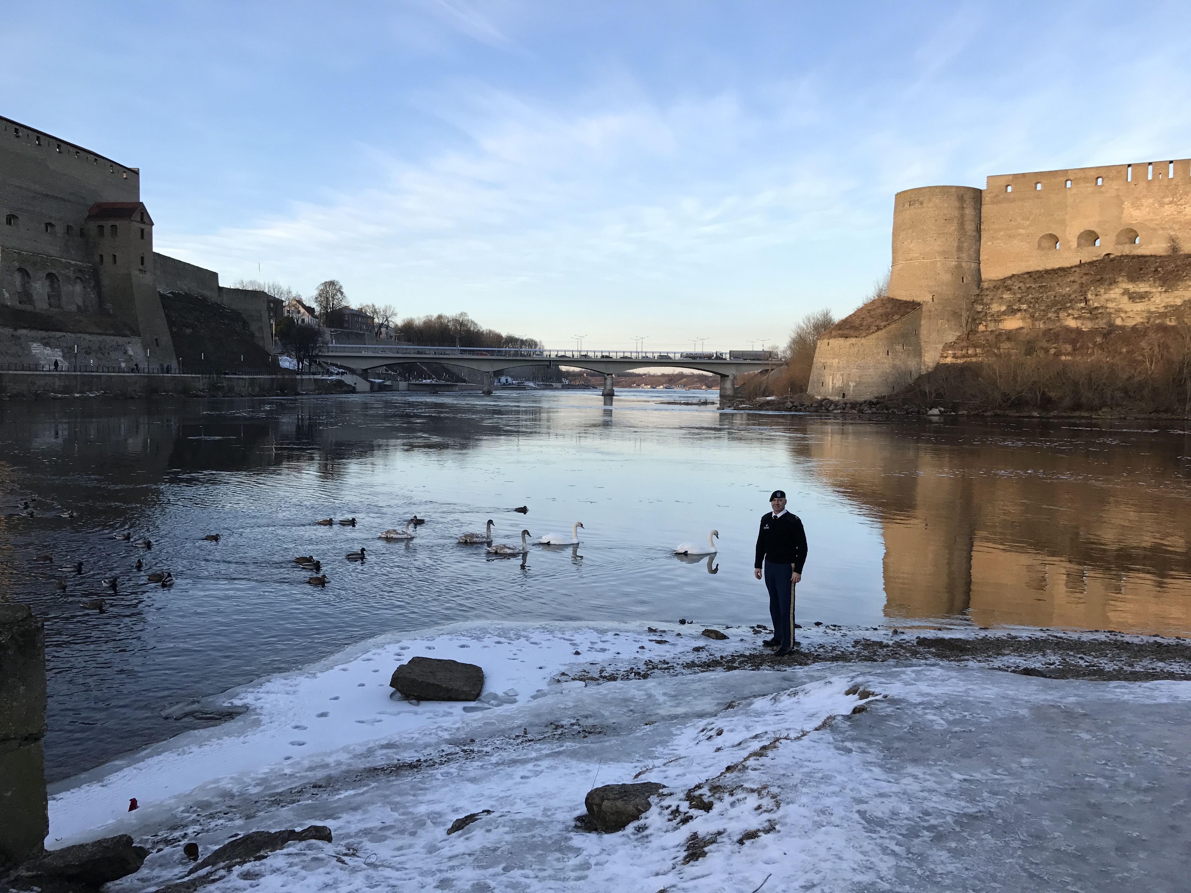 Senator Mastriano standing along in uniform along an icy river between Estonia and Russia. The sky is a beautiful blue and there are buildings on either side of him.
