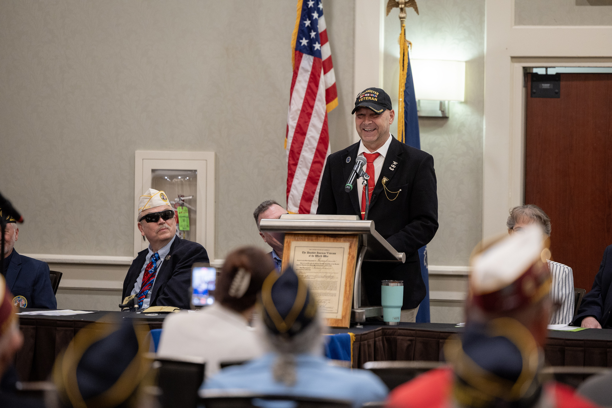 Senator Mastriano standing in front of Veterans speaking in a black sports jacket and red tie and a ballcap