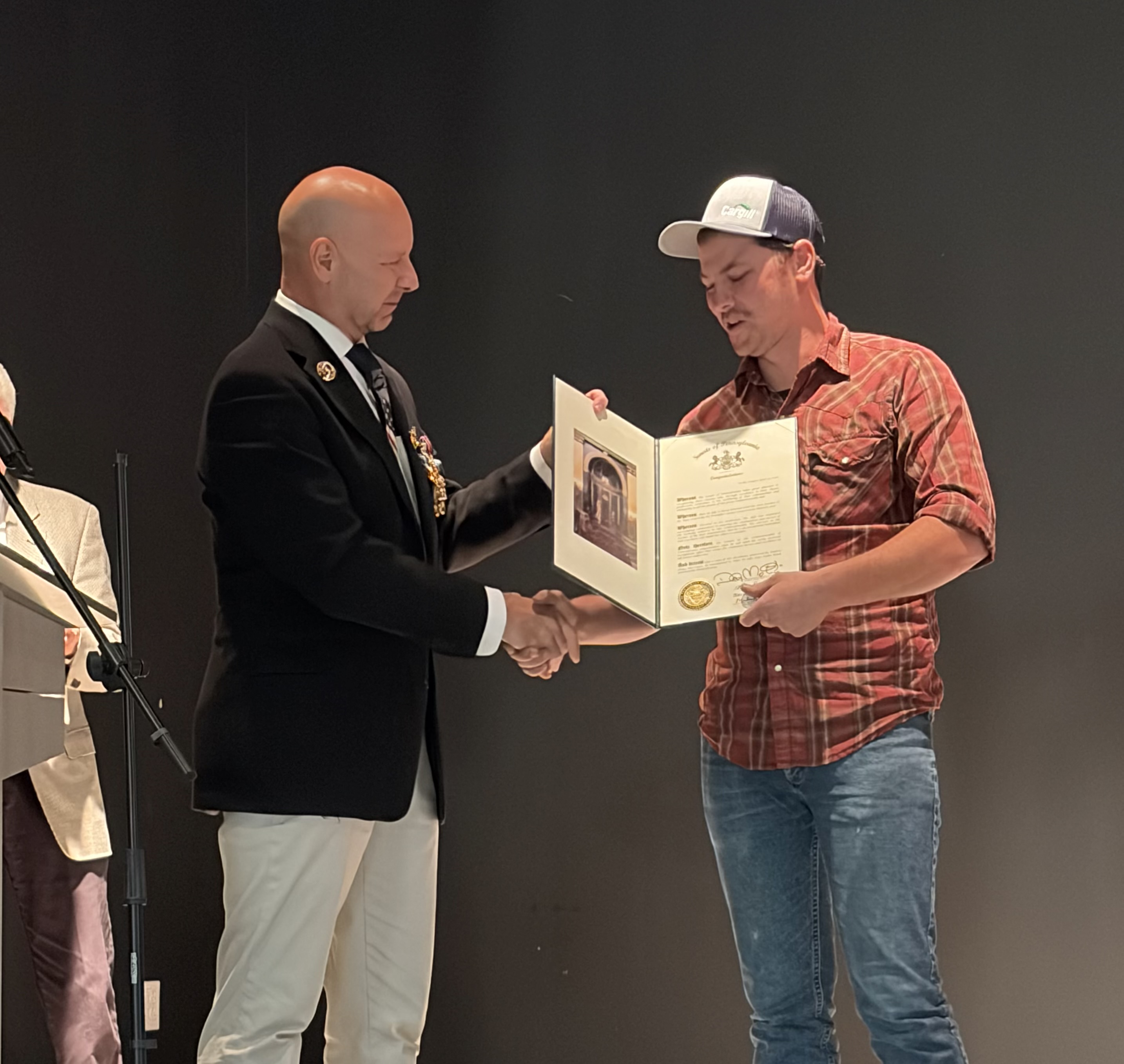 Senator Mastriano in a black sports jacket and beige pants gives a senate citation to a gentlemen wearing a rust colored plaid shirt, hat and jeans