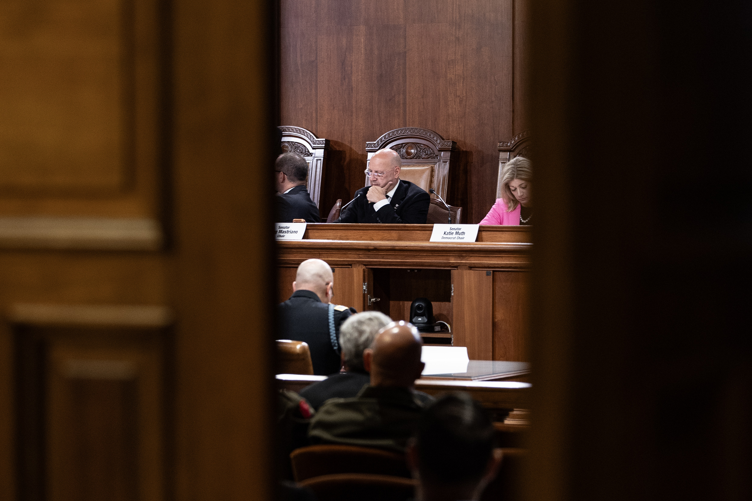 View through a doorway into a formal hearing room where officials sit at a wooden dais, with audience members in the background.
