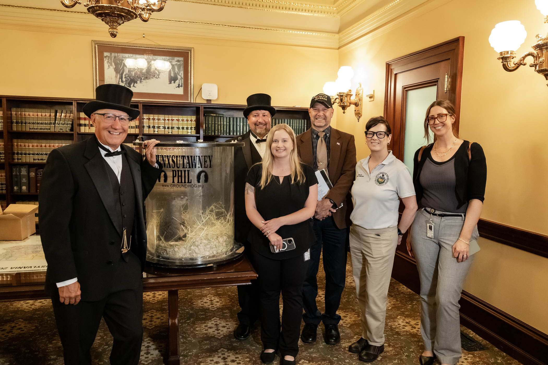 6 people standing in a room at the PA State Capitol with Punxsutawney Phil. Senator Mastriano is in the middle wearing a ballcap, sports jacket and tie. He has three female staff next to him.