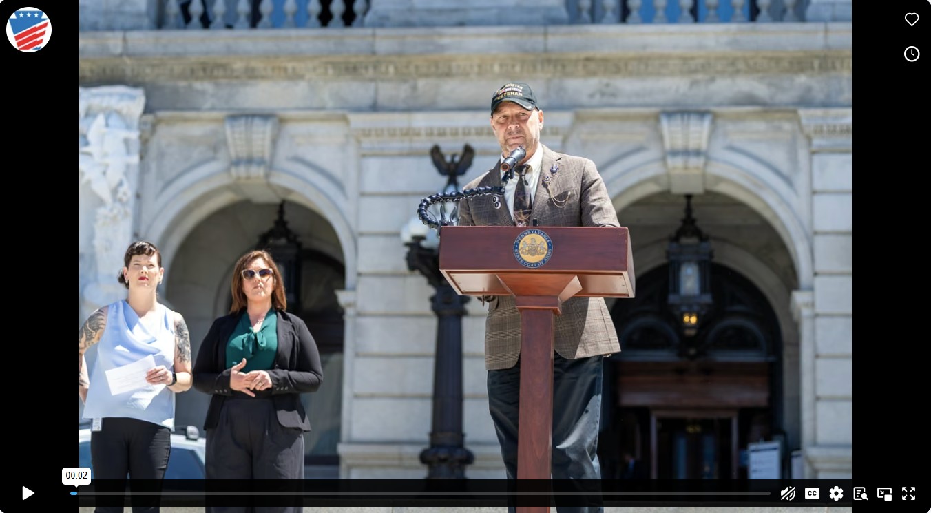 Senator Mastriano wearing a tan plaid blazer, Afghanistan Veteran ball cap standing at the podium on the Harrisburg State Capitol steps speaking about Jonny Tomasello