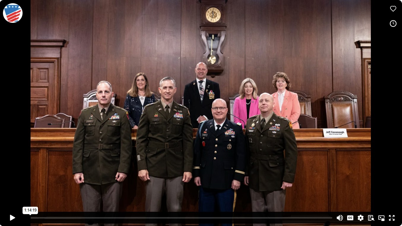 Group photo in a wood paneled hearing room showing Senator Mastriano centered in the back row above a dais, with uniformed military officers standing in front.