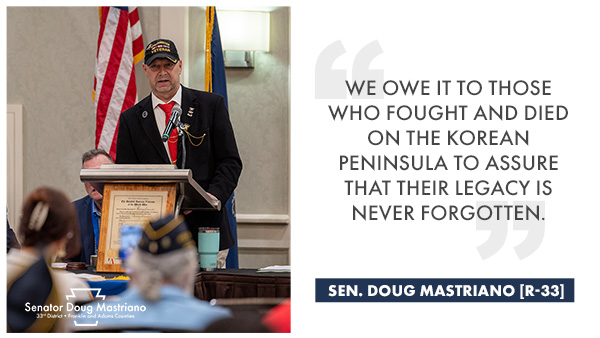 Senator Mastriano stands at a podium in a black sports jacket, red tie, white shirt, veterans ball cap. Both the American flag and PA state flag are behind him