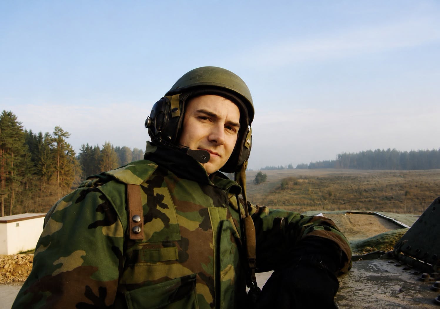 Senator Mastriano in green, brown and black Army gear standing against a dark metal container. Blue skies and trees in the background.