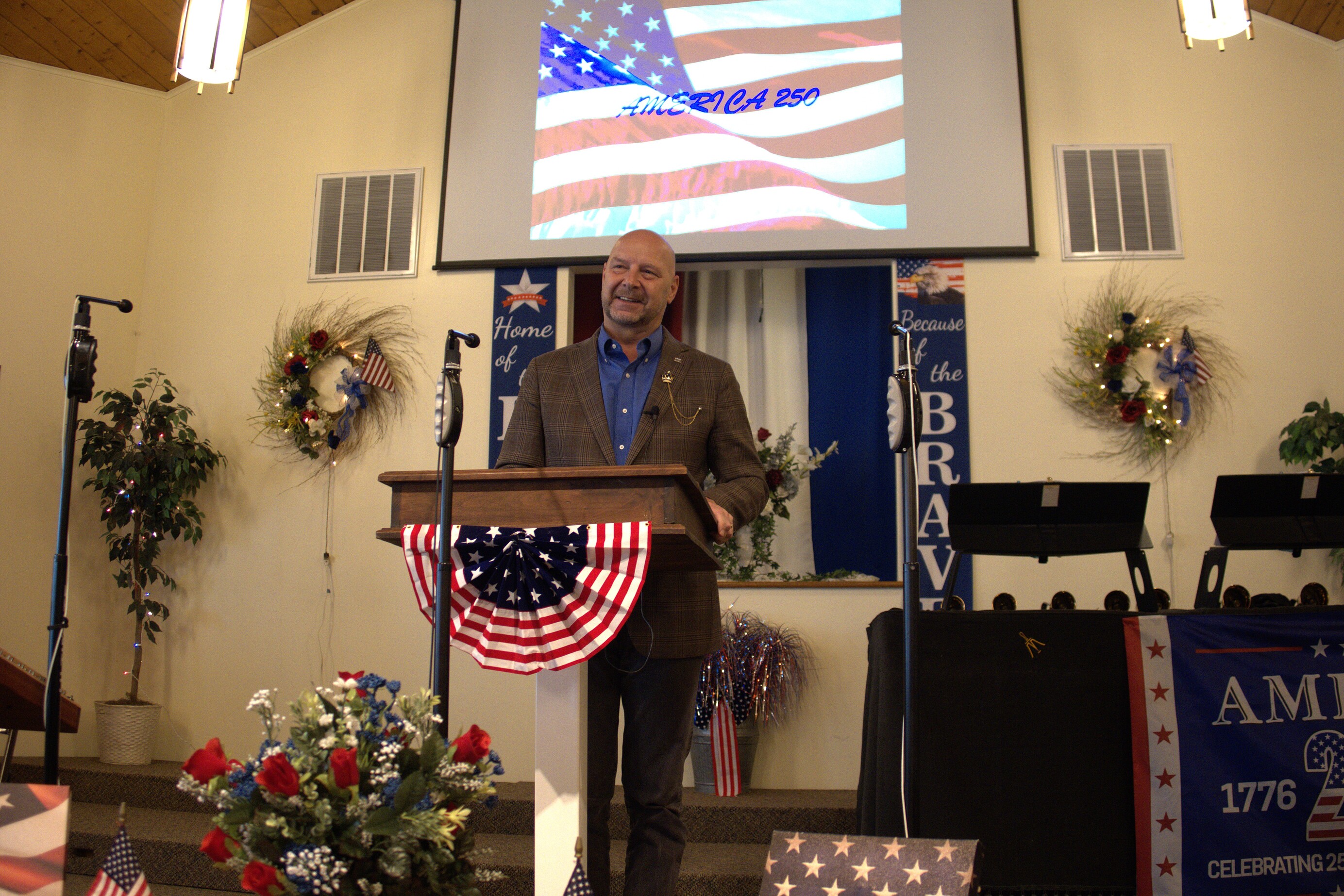 Senator Mastriano wearing a brown plaid sports jacket, blue dress shirt, standing at a podium in front of a church with the American flag on a screen back behind him. There is also flag décor on the outside of the podium