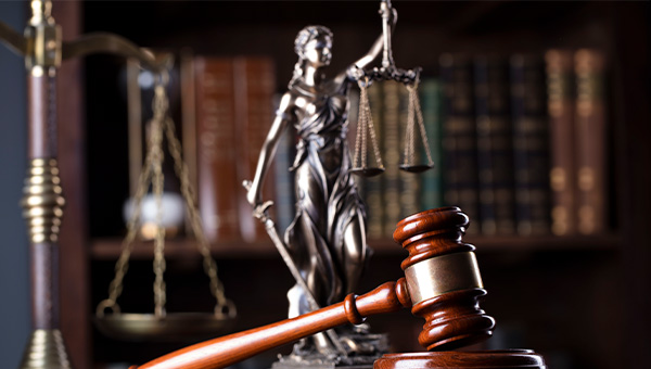 Courtroom gavel displayed in front of “Blind Justice” statuette holding up scales of justice, with law library books in background