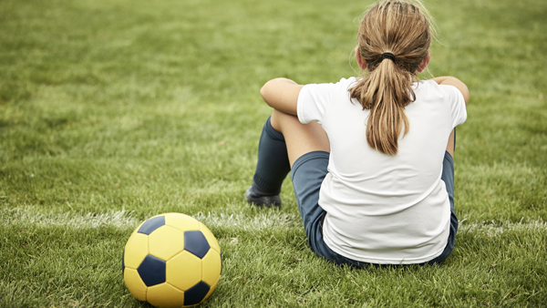 Girl sits on the edge of a grass soccer field with yellow soccer ball by her side.