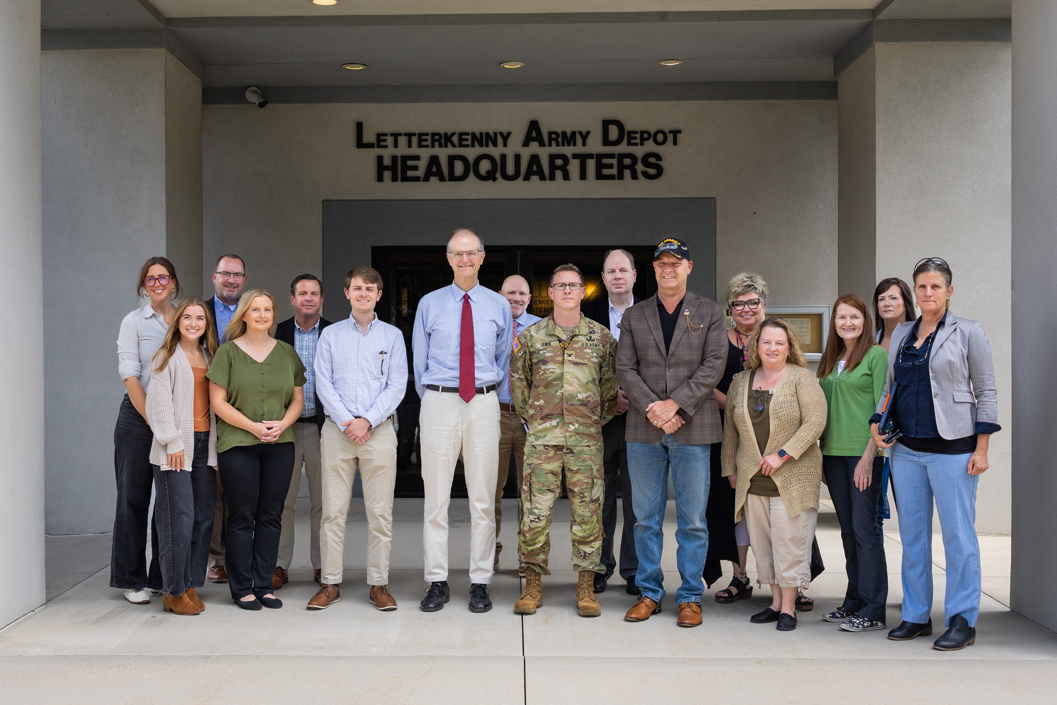 large group of approximately fifteen professionally dressed individuals stands smiling together for a group photograph in front of the entrance to the Letterkenny Army Depot Headquarters. The group stands on a concrete landing between two large pillars under an overhanging roof. The entrance behind them features large dark lettering above the doorway that reads "LETTERKENNY ARMY DEPOT HEADQUARTERS." The individuals are in a mix of professional attire—including button-down shirts, slacks, blazers, and dresses—with one central figure dressed in a military camouflage uniform and Senator Mastriano wearing a veteran's cap.
