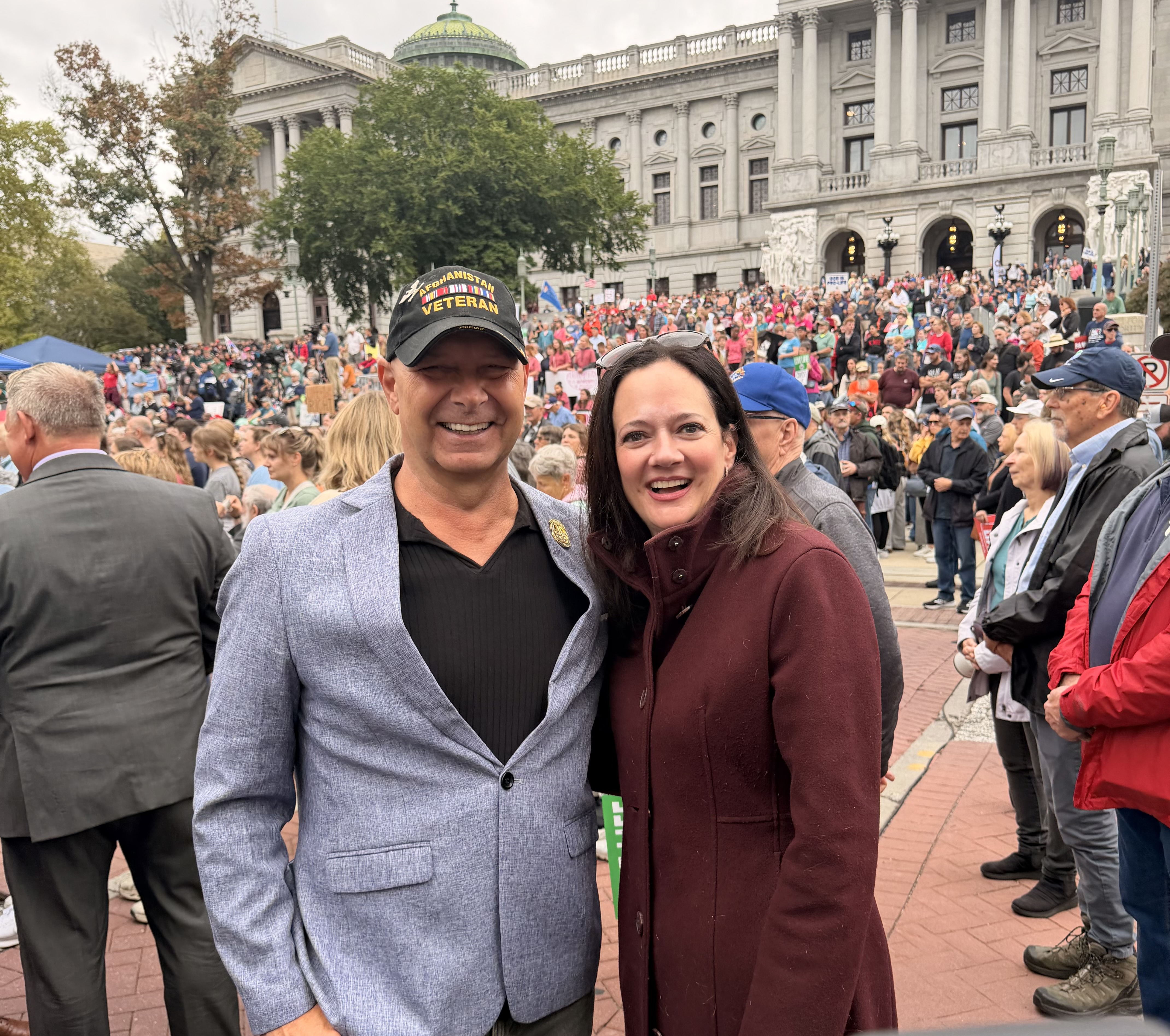 Senator Mastriano wearing a light blue sports jacket, black shirt and black hat. Next to him, Representative Borowicz is wearing a maroon coat standing in front of the Harrisburg Capitol with thousands of people behind them.