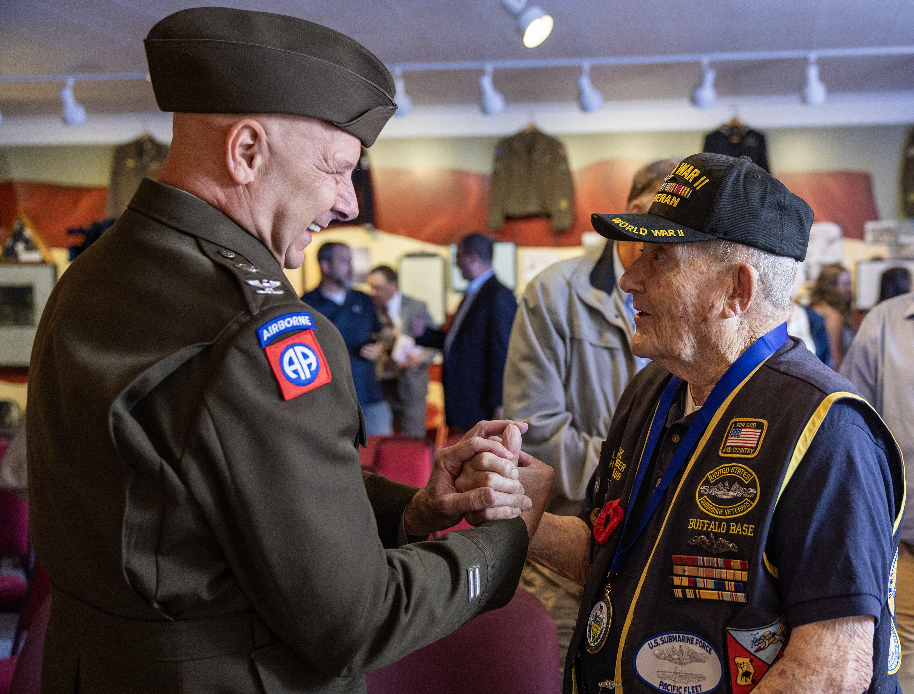 A side-profile photograph of Pennsylvania Senator Doug Mastriano wearing a military uniform, smiling warmly as he firmly shakes hands with an elderly World War II veteran. Senator Mastriano is in an Army service uniform featuring an "Airborne" and "82nd Airborne Division" patch on his left shoulder. The veteran wears a black "World War II Veteran" cap and a navy blue vest heavily adorned with military patches, ribbons, and a large circular emblem that reads "Buffalo Base." They are standing indoors at an event or museum space with other people and military uniform displays blurred in the background.