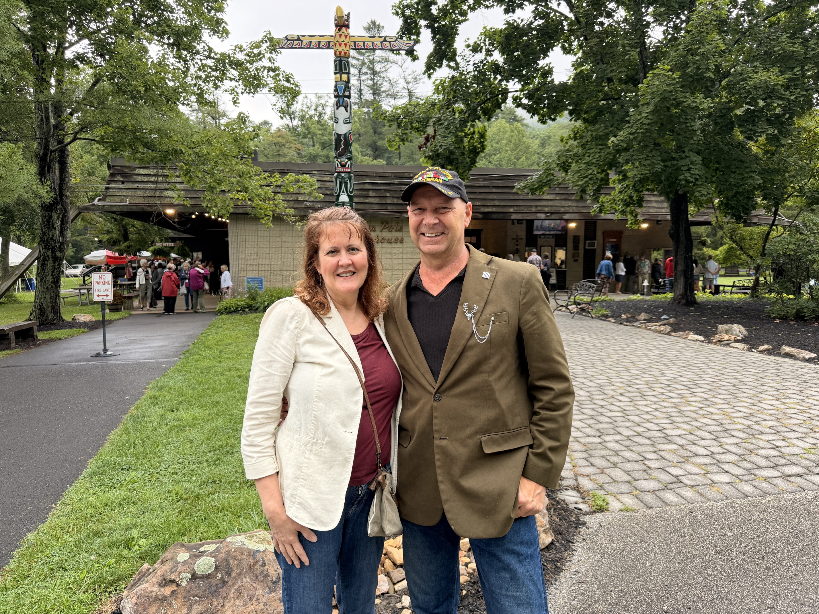 Pennsylvania State Senator Doug Mastriano stands smiling next to his wife, Rebbie Mastriano, outside the Totem Pole Playhouse in Fayetteville, Pennsylvania. Senator Mastriano wears a brown blazer over a black V-neck shirt, blue jeans, and a camouflage baseball cap, while his wife wears a white open blazer over a maroon shirt and blue jeans. They are standing on a gravel walkway with a green lawn to their left and thick green trees behind them. In the background, the rustic wooden building of the playhouse features its signature tall totem pole rising high above the roofline under an overcast sky.
