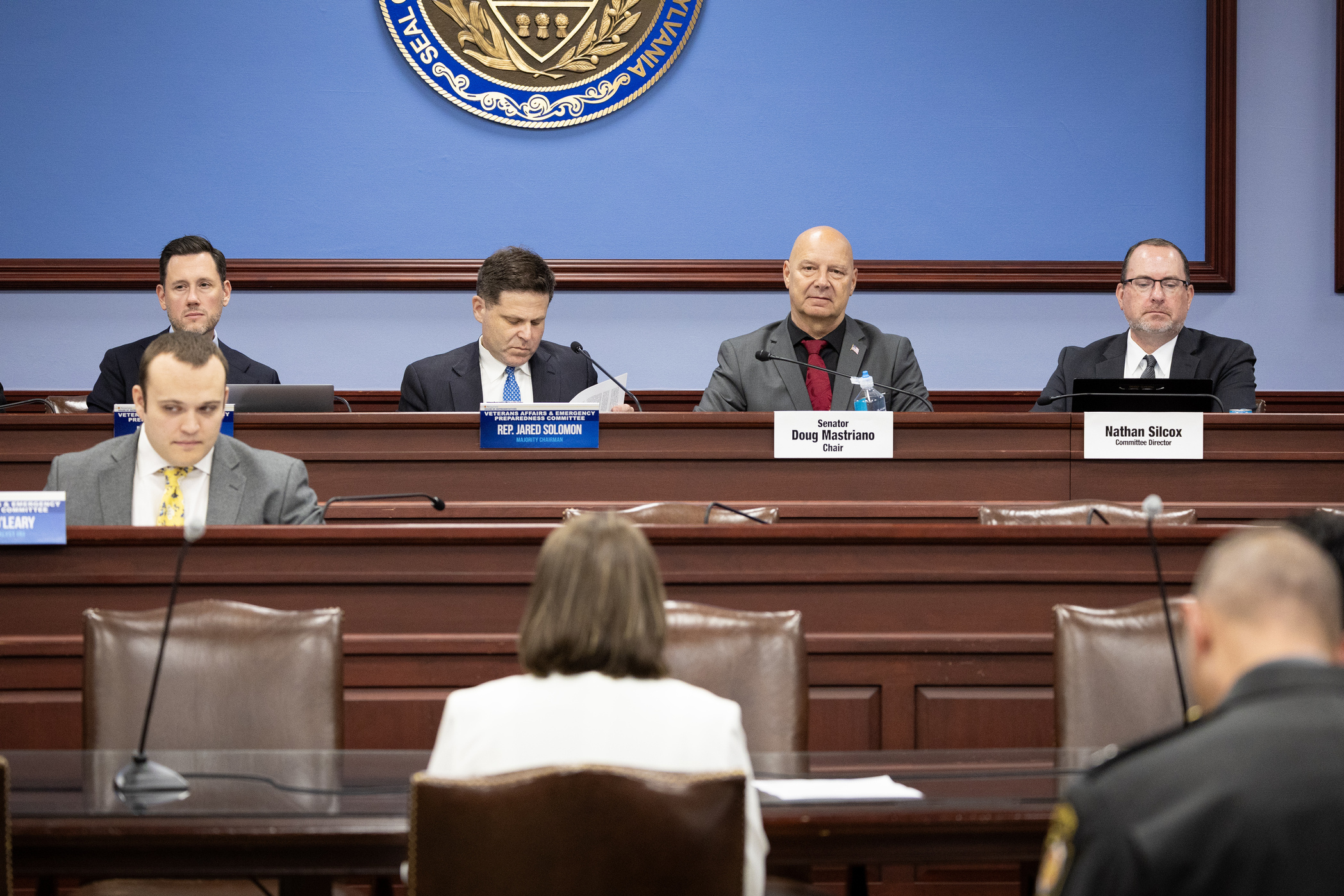 A state committee hearing takes place inside a formal room with wood paneling and a blue background featuring the Pennsylvania state seal. Four committee members sit at a raised dais, looking down at an individual seated in front of them with their back to the camera. The panel includes a nameplate for Senator Mastriano, designated as Chair, alongside plates for Rep. Jared Solomon and Nathan Silcox, the Committee Director. Microphones are positioned on the desks, and papers are scattered before the officials as they conduct the proceedings.