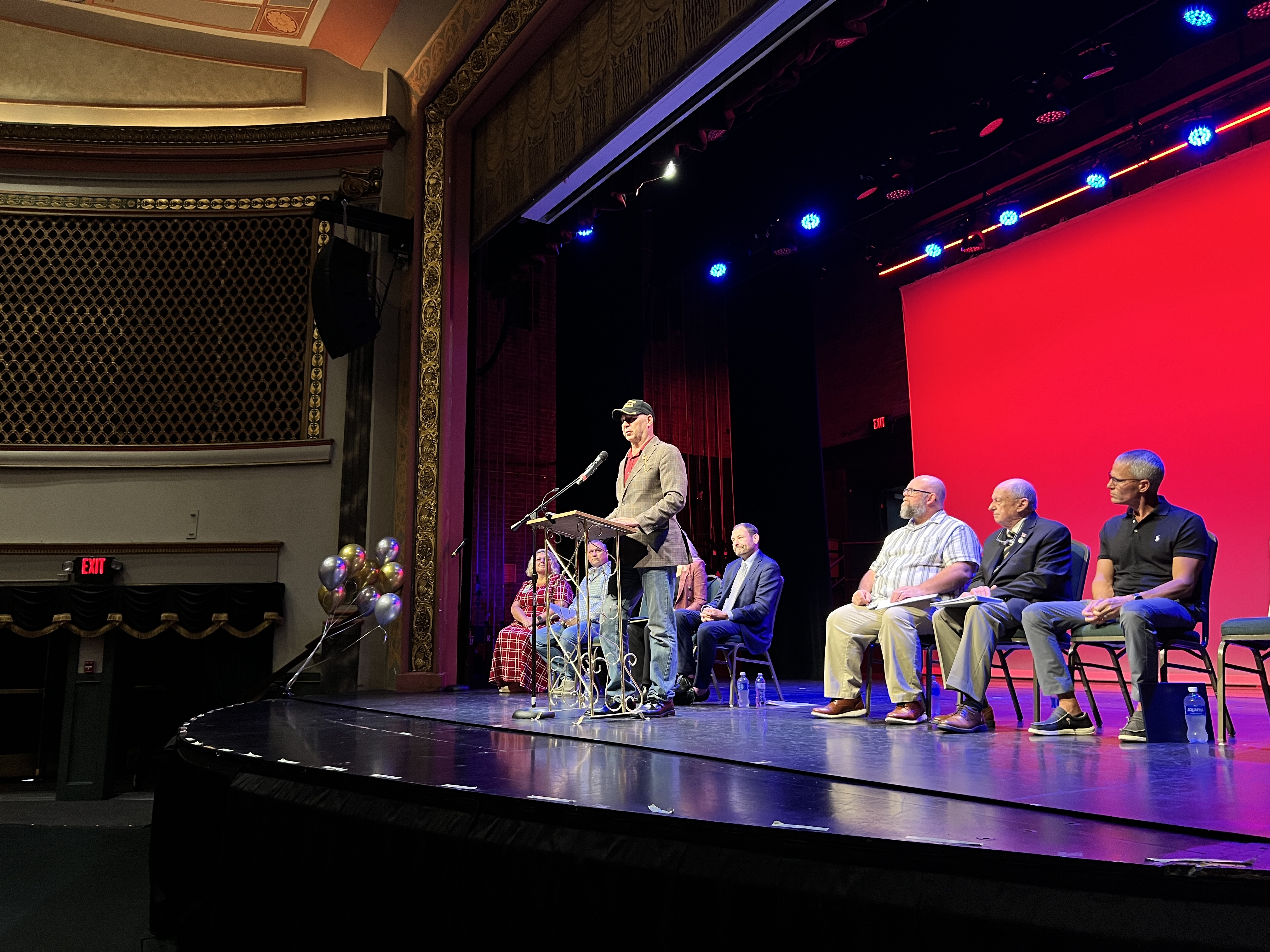 Senator Doug Mastriano speaks from a metal podium on an illuminated theater stage. He is wearing a dark baseball cap, sunglasses, a red shirt, a tan blazer, and blue jeans. Behind him, several local officials sit in a row of chairs on the stage, listening intently. The background features a large, bright red screen lit by overhead blue spotlights. The ornate historical architecture of the theater's balcony and proscenium arch is visible on the left.