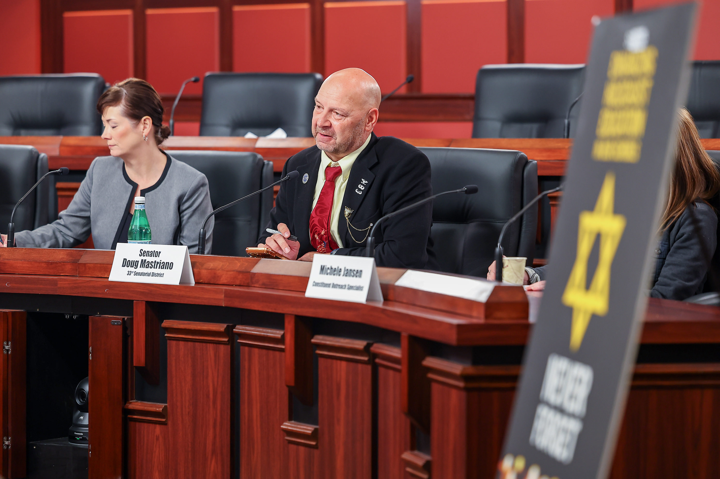 Senator Doug Mastriano sits at a curved wooden committee bench inside a legislative chamber. He is wearing a black suit jacket, a pale yellow shirt, and a red patterned tie while holding a pen. Next to him is a female colleague, and nameplates for both individuals sit on the desk in front of them. In the foreground, a partially blurred banner features a Star of David and the words "NEVER FORGET