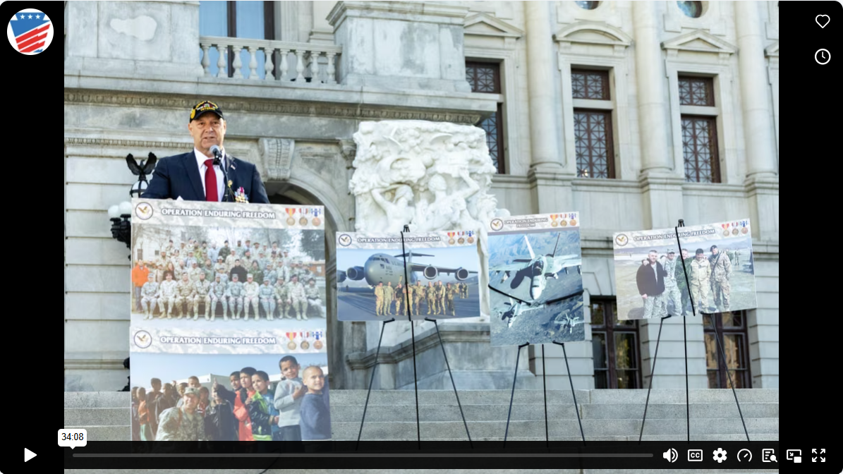 Senator Mastriano speaking at a podium on stone steps in front of a building, flanked by display boards labeled "Operation Enduring Freedom."