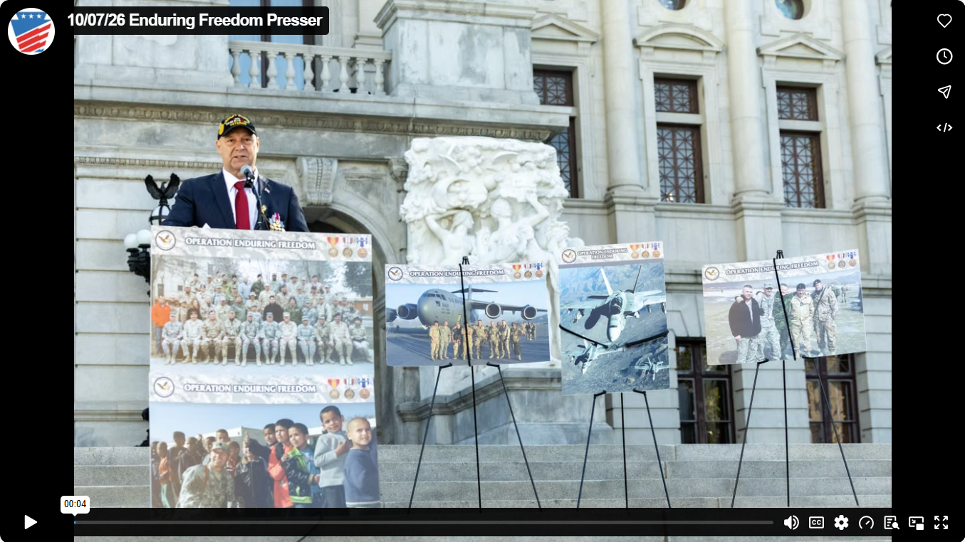 Senator Mastriano speaking at a podium on stone steps in front of a building, flanked by display boards labeled "Operation Enduring Freedom."