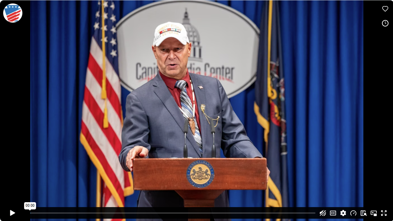 Senator Doug Mastriano wears a suit and veteran cap while speaking at a Capitol Media Center podium flanked by American and Pennsylvania flags.