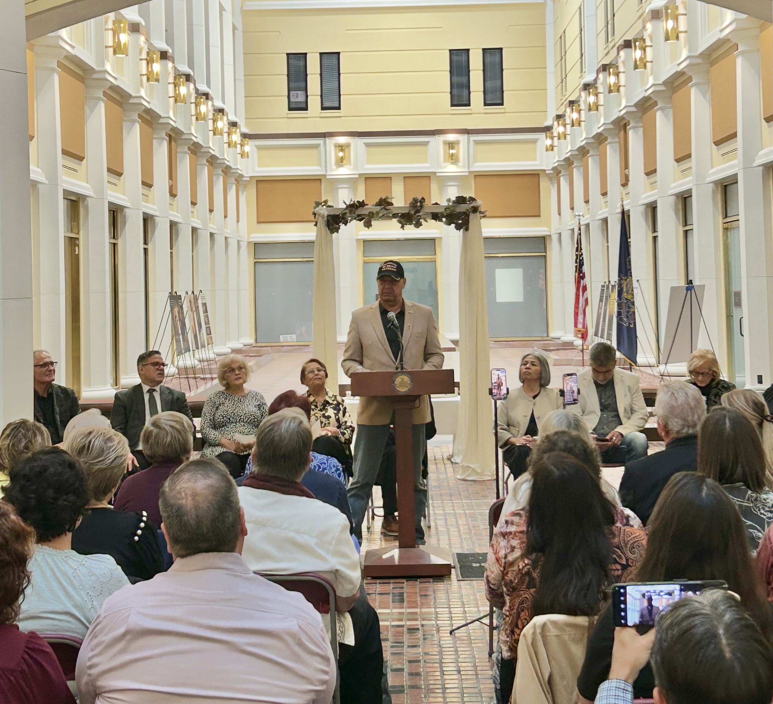 An indoor photograph captures Pennsylvania State Senator Doug Mastriano speaking from a wooden podium at the Pennsylvania State Capitol. He is wearing a tan blazer, blue jeans, and a black baseball cap that reads "VETERAN." There are many people in the crowd sitting while he speaks.