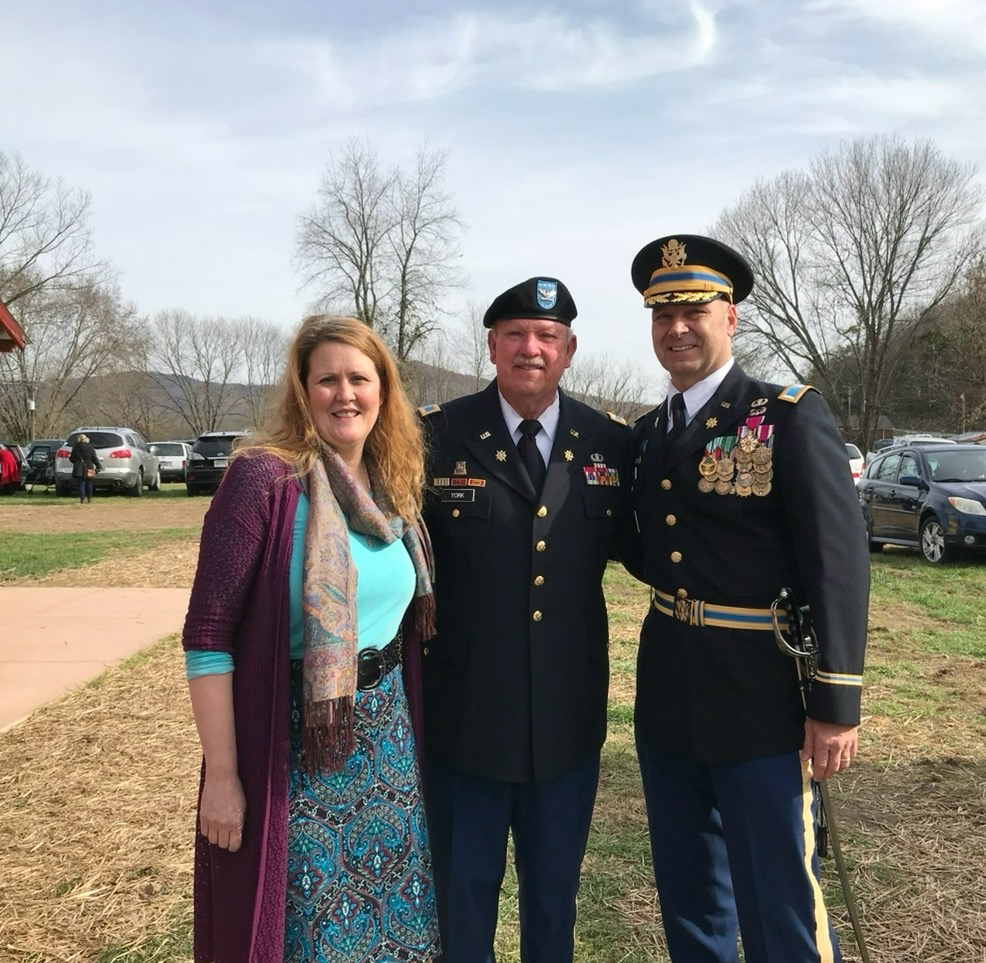 Senator Doug Mastriano in military dress uniform and Rebbie Mastriano smile with a serviceman outdoors on a sunny day with trees in the background.