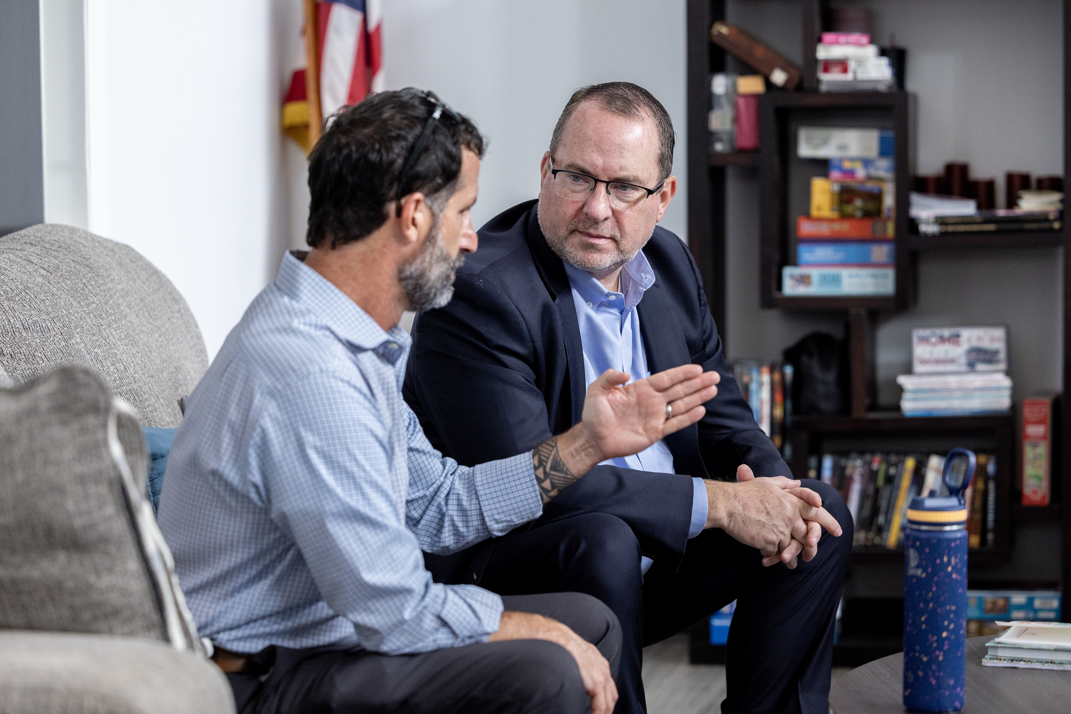 Two men are seated indoors engaged in a conversation. On the left, a man in a light blue patterned button-down shirt and grey trousers sits on a couch, gesturing with his left hand, which reveals a geometric tattoo on his forearm. On the right, a man with glasses, wearing a navy-blue blazer and a light blue dress shirt, sits opposite him, listening intently with his hands clasped. The background includes a bookshelf filled with items and a partially visible American flag.