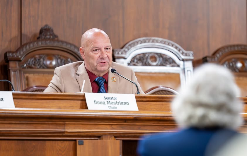 Pennsylvania State Senator Doug Mastriano sits at a legislative committee bench, speaking behind a microphone with a nameplate reading "Senator Doug Mastriano Chair" visible in front of him.